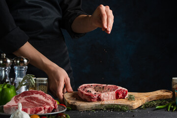 Close-up view of chef pours pepper on raw steak on wooden chopped board. Backstage of preparing grilled pork meat at restaurant kitchen on dark blue background. Frozen motion. Cooking process.