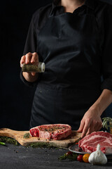 Chef pours spices on raw steak on wooden chopped board. Backstage of preparing grilled pork meat at restaurant kitchen on dark blue background. Frozen motion. Cooking process.
