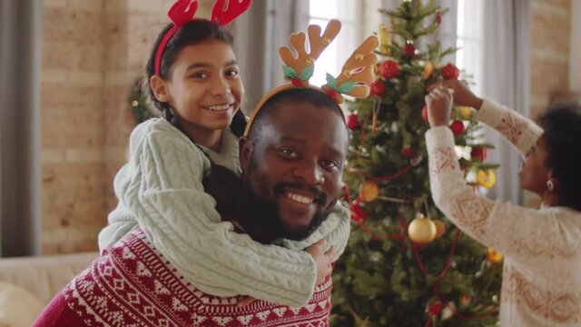 Portrait of happy Afro-American father in deer antler headband holding cheerful daughter on his back, looking at camera and smiling while mother decorating Christmas tree in background