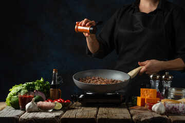 Professional chef in black uniform pours fragrant spices into pan with meat filling for cooking tacos on dark blue background. Traditional mexican cuisine. Concept of tasty street food. Frozen motion.