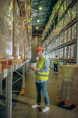 Young warehouse worker in helmet standing near containers and holding a tablet
