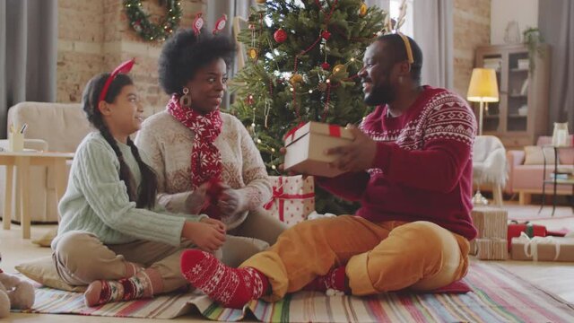 Happy Afro-American man receiving Christmas present from loving wife and daughter, opening box, taking off woolen scarf, saying thanks and giving hugs to them while sitting on floor in living room
