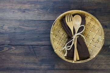 Wooden set spoon and fork with brown napkin put on bamboo woven plate. Rustic wooden background, top view, copy space for text.