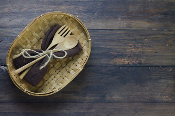 Wooden set spoon and fork with brown napkin put on bamboo woven plate. Rustic wooden background, top view, copy space for text.