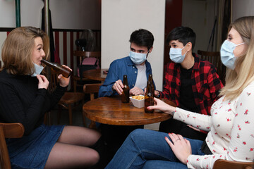 Four friends wearing face masks in a pub. One of them is drinking and other are looking at her. New...