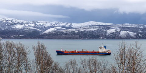 Cargo ship sails along the sea Bay against the background of snow-capped mountains. Cold autumn weather. Sea freight and shipping. Nagaeva Bay, sea of Okhotsk, Magadan region, Far East of Russia.