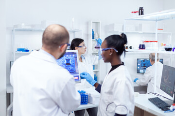 Scientist holding glass flask with blue genetic material in busy laboratory. Multiethnic team of medical researchers working together in sterile lab wearing protection glasses and gloves.