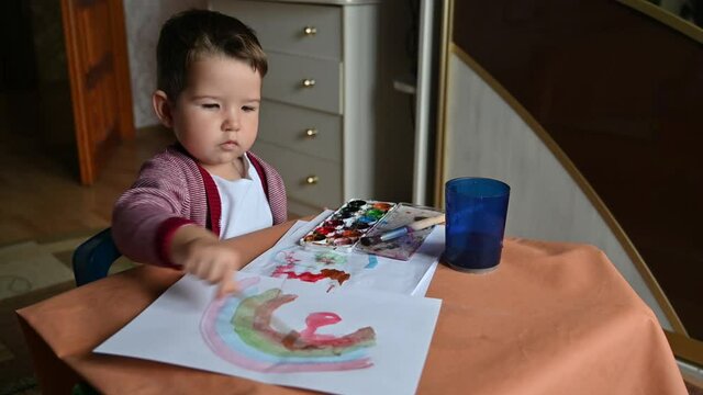 A 2 years old child draws a rainbow with paints. Creative activities at home concept. Children's desk, drawing supplies.
