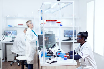 Fototapeta na wymiar Scientist with african ethnicity holding sample recipient working with her team in modern facility. Black healthcare researcher in biochemistry laboratory wearing sterile equipment.