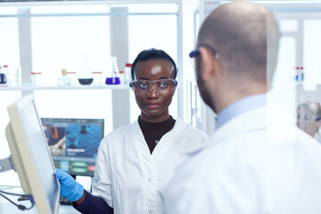 Fototapeta premium African scientist discussing virus trial with virolog in chemistry laboratory. Multiethnic team of medical researchers working together in sterile lab wearing protection glasses and gloves.