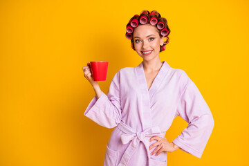 Portrait of attractive cheerful woman wearing curlers bathrobe drinking tea isolated over shine yellow color background