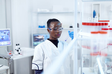 African scientist in protective glasses looking at test tube with genetic material. Black researcher in sterile laboratory conducting pharmacology experiment wearing coat.