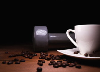 Heavy dumbbell and small cup of hot coffee. Fresh unground coffee beans scattered on a wooden table, with black background. Healthy fitness lifestyle concept.