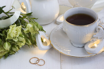 Obraz na płótnie Canvas a white porcelain coffee cup on a saucer with wedding rings lying next to it, white bush roses and heart-shaped lights on a white wooden background