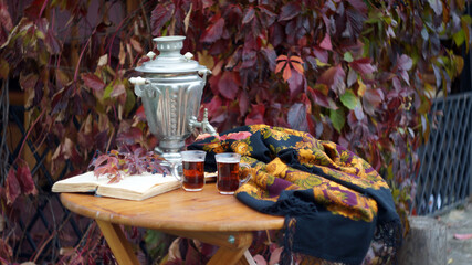round wooden table, standing on it a samovar, glasses of tea, lying next to a book and a bright shawl with a yellow floral pattern on a background of purple autumn leaves.  autumn still life.   