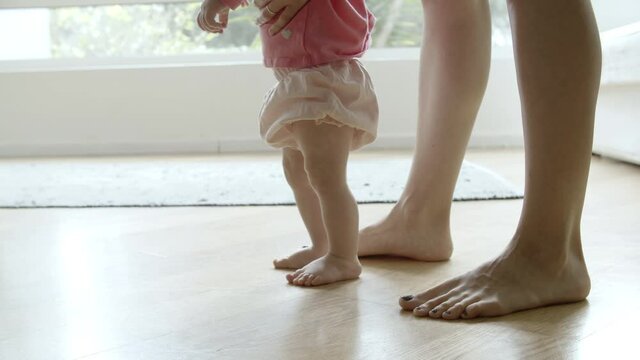 Cute barefoot baby learning walking with help of mother on floor. Side view of Caucasian loving mom legs going behind little baby and holding son or daughter. Making first step and family concept