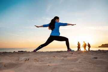 Fototapeta premium A young woman in sportswear trains by performing a leg stretch. In the background, sunset, sandy beach and ocean. Rear view. Wellness and sports lifestyle