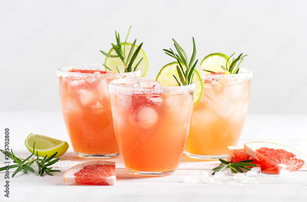 Wall mural Three glasses of paloma cocktail with ice and tequila, decorated with lime wedges, grapefruit and rosemary stand on a white wooden table. Horizontal orientation, selective focus