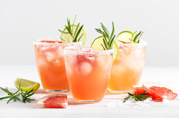 Three glasses of paloma cocktail with ice and tequila, decorated with lime wedges, grapefruit and rosemary stand on a white wooden table. Horizontal orientation, selective focus
