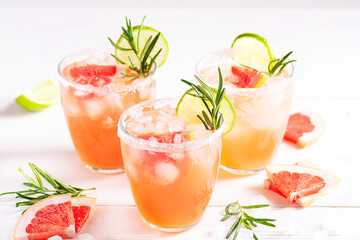 Paloma cocktail with ice and tequila, garnished with lime wedges, grapefruit and rosemary on a white wooden table. Horizontal orientation, selective focus