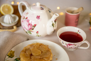 Cute porcelain teapot and teacup and a mini tarte Tatin on an elegant table with white table cloth, sugar and lemon for the tea in the background.