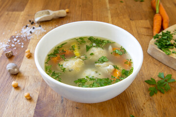 White bowl of chicken soup with chopped parsley on top, on a butcher's block surface, with some salt and peppers, carrots and parsley. 