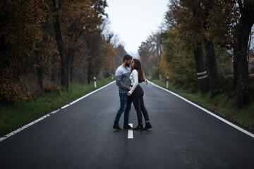 Beautiful portrait of young couple hugging and standing on a perfect clean long road bewtween the green forest in autumn