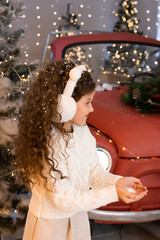 little girl playing with snow near red car and Christmas trees with lights. Merry Christmas and Happy Holidays