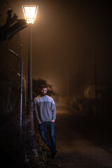 Young good-looking man with black beard leaning on a street light at night time under its light