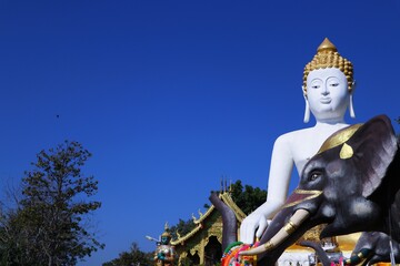 Fototapeta premium White Buddha statue at Wat Phra That Doi Kham in Chiang Mai, Thailand, under a clear blue sky, representing Buddhist spirituality, peace, faith, meditation, and northern Thai cultural heritage.