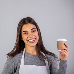 Happy brunette waitress in apron offering you glass with hot coffee while standing in front of camera in isolation. A waitress holding and serving a paper cup of hot coffee