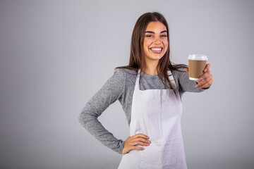 Happy brunette waitress in apron offering you glass with hot coffee while standing in front of camera in isolation. Business Owner Concept - Beautiful Caucasian Barista offers disposable take away