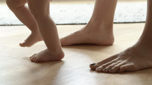 Barefoot infant learning walking with help of mother on floor. Closeup side view of Caucasian mom feet going behind little baby and holding son or daughter. Making first step and family concept