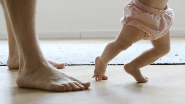Barefoot infant learning walking with help of father. Side closeup view of Caucasian dad legs walking behind little baby and holding son or daughter. Making first step and family concept