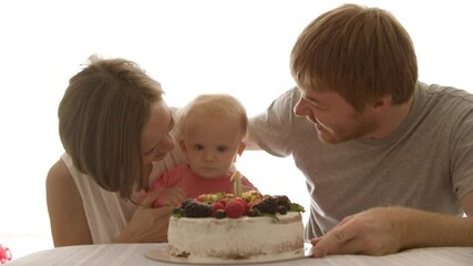 Young parents blowing candle on cake with baby girl and clapping. Happy Caucasian mom and dad celebrating first birthday of cute little daughter in pink shirt at home. Celebration and family concept