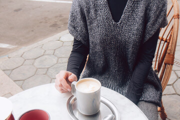 Woman in a cozy gray-blue vest spends time outdoors in the city. Female sits in street cafe on a wooden wicker chair in street cafe. Cup of morning coffee. Noface