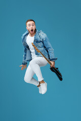 attractive crazy man in denim t-shirt jeans with guitar jumping high isolated on blue background