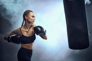 Naklejka premium sporty brunette woman in boxing gloves and sportswear kicking bag on dark background with smoke.