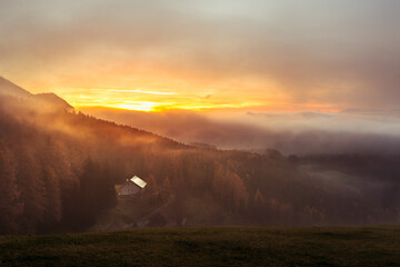 Sonnenuntergang auf der Alm wo ein Bauernhof zu sehen ist links unten umgeben von Nebel und Wald