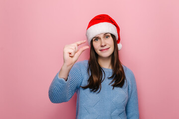 Young woman shapes little gesture, demonstrates something very tiny, wears winter sweater and Christmas red hat, measures small size, isolated over pink background with copy space. Not very much