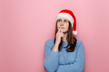 Confused young female in Christmas hat with puzzled expression, spreads hands with confused look, stands puzzled, isolated on pink studio background wall. Happy New Year celebration merry holiday