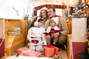 Happy parents and children with gift boxes in their hands in red car in pickup trailer near Christmas tree. © Marharyta