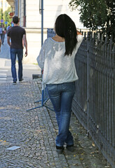 Full-length portrait of a young brunette in blue jeans and a white knitted blouse on a city street near a cast-iron fence. View from the back.