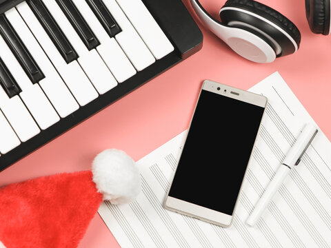 Mobile Phone , Blank Music Sheet , Christmas Santa Hat, Headphones And  Electric Piano On Pink Background With Copy Space.Remote Piano Lessons And Christmas Party.