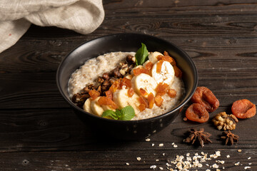 Oatmeal with dried apricots, banana and walnuts, cooked in milk and decorated with mint chanterelles. Healthy diet vegetarian breakfast in a round ceramic bowl on a wooden kitchen table.