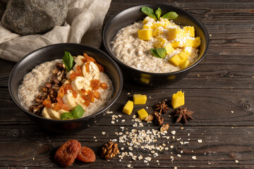 Oatmeal with fresh mango pieces, banana, dried apricots and walnuts, cooked in milk and decorated with mint chanterelles. Two servings of classic vegetarian breakfast on a wooden kitchen table.