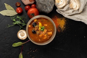 A bowl of spicy tomato soup with meat, olives and mushrooms on a black stone kitchen table.