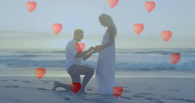 Multiple heart balloons floating against man proposing a woman with a ring on beach
