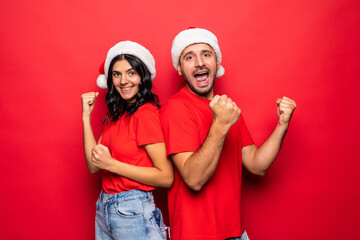 Fototapeta premium Cheerful young couple with win gesture in Santa Claus hats together isolated over red background