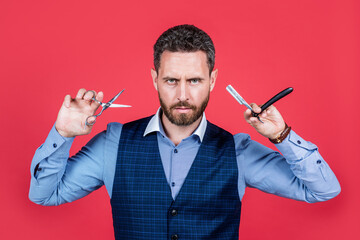 Handsome man with unshaven facial hair hold scissors and open razor vintage barber tools red background, barbershop